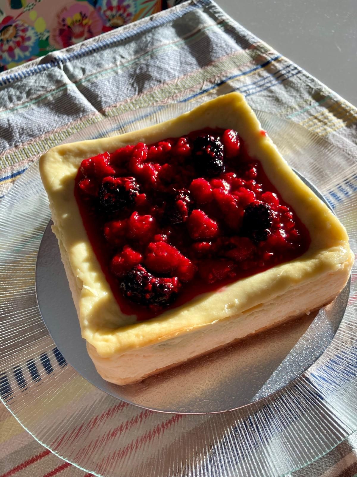 Square cheesecake topped with a vibrant berry compote, displayed on a textured tablecloth under sunlight.