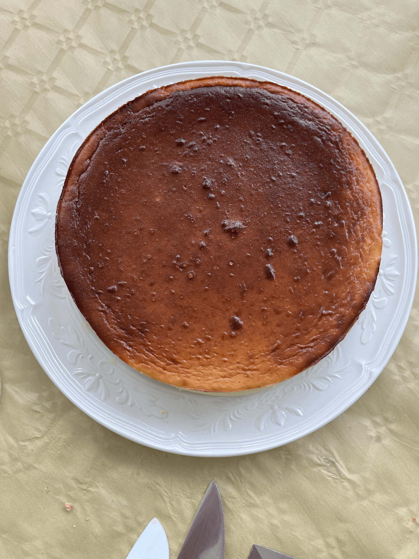 Whole cheesecake on a decorative white plate, placed on a patterned tablecloth with a knife nearby.