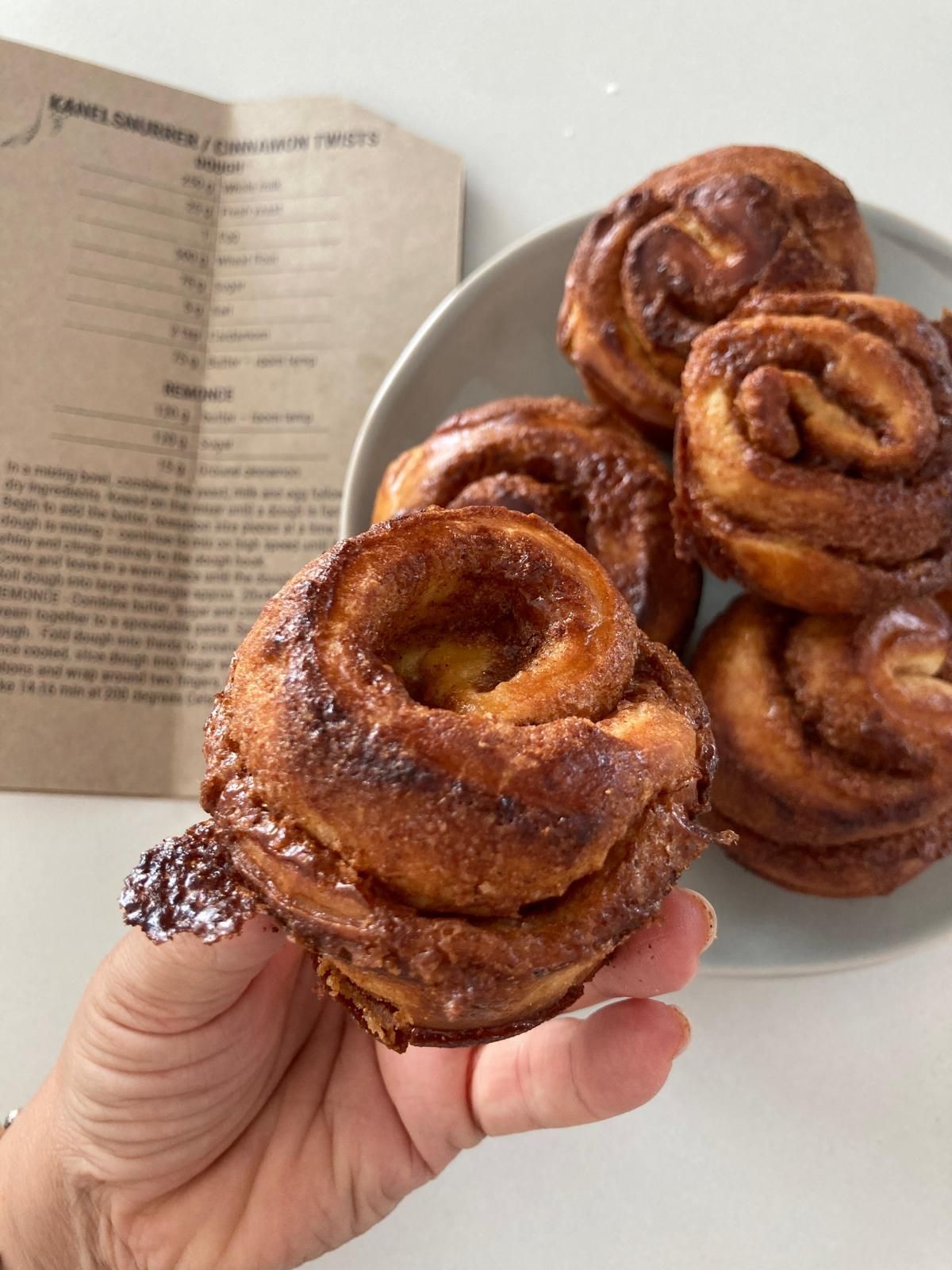 Hand holding a cinnamon roll with more rolls on a plate, recipe book in the background.