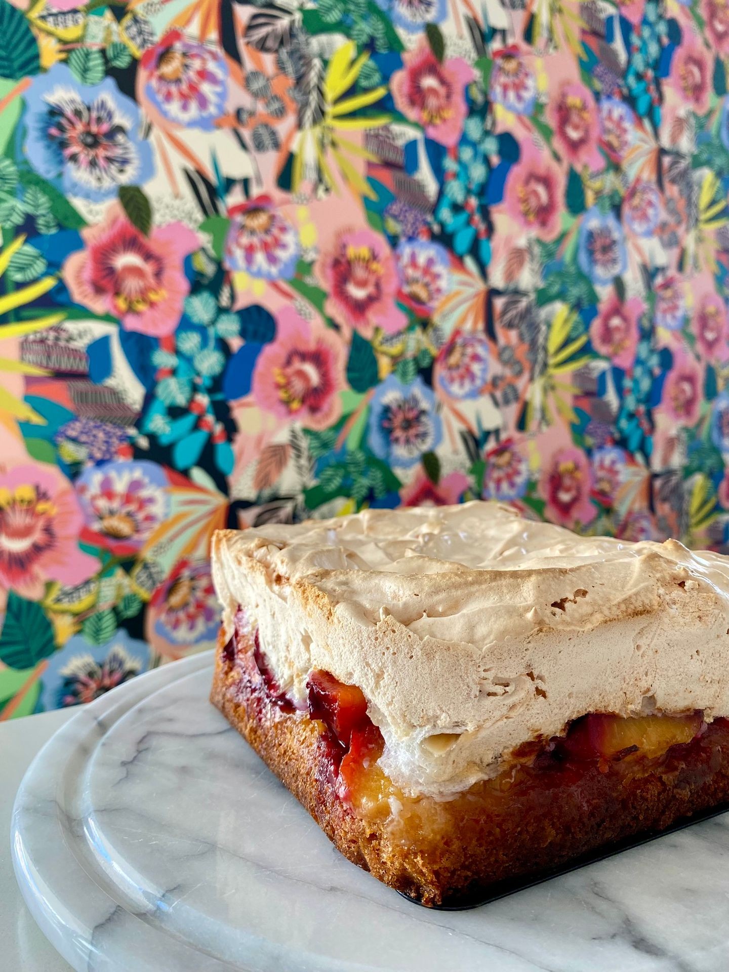 Square meringue-topped dessert on a marble platter with vibrant floral wallpaper background.