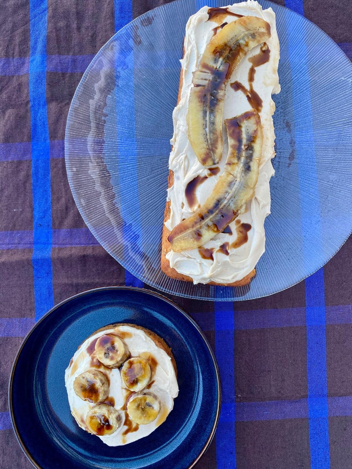 Cake with sliced bananas and cream on a glass and blue plate on a checkered tablecloth.