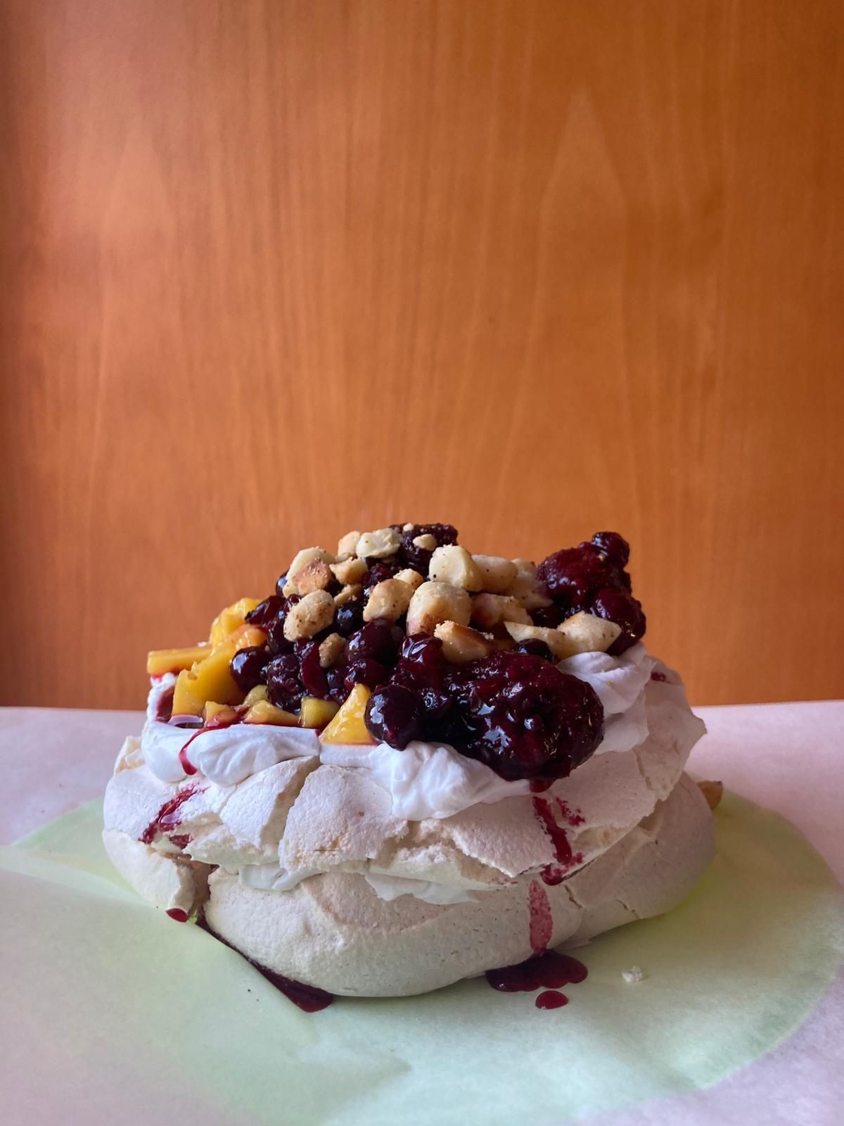 Pavlova topped with whipped cream, berries, and mango on a light green surface.