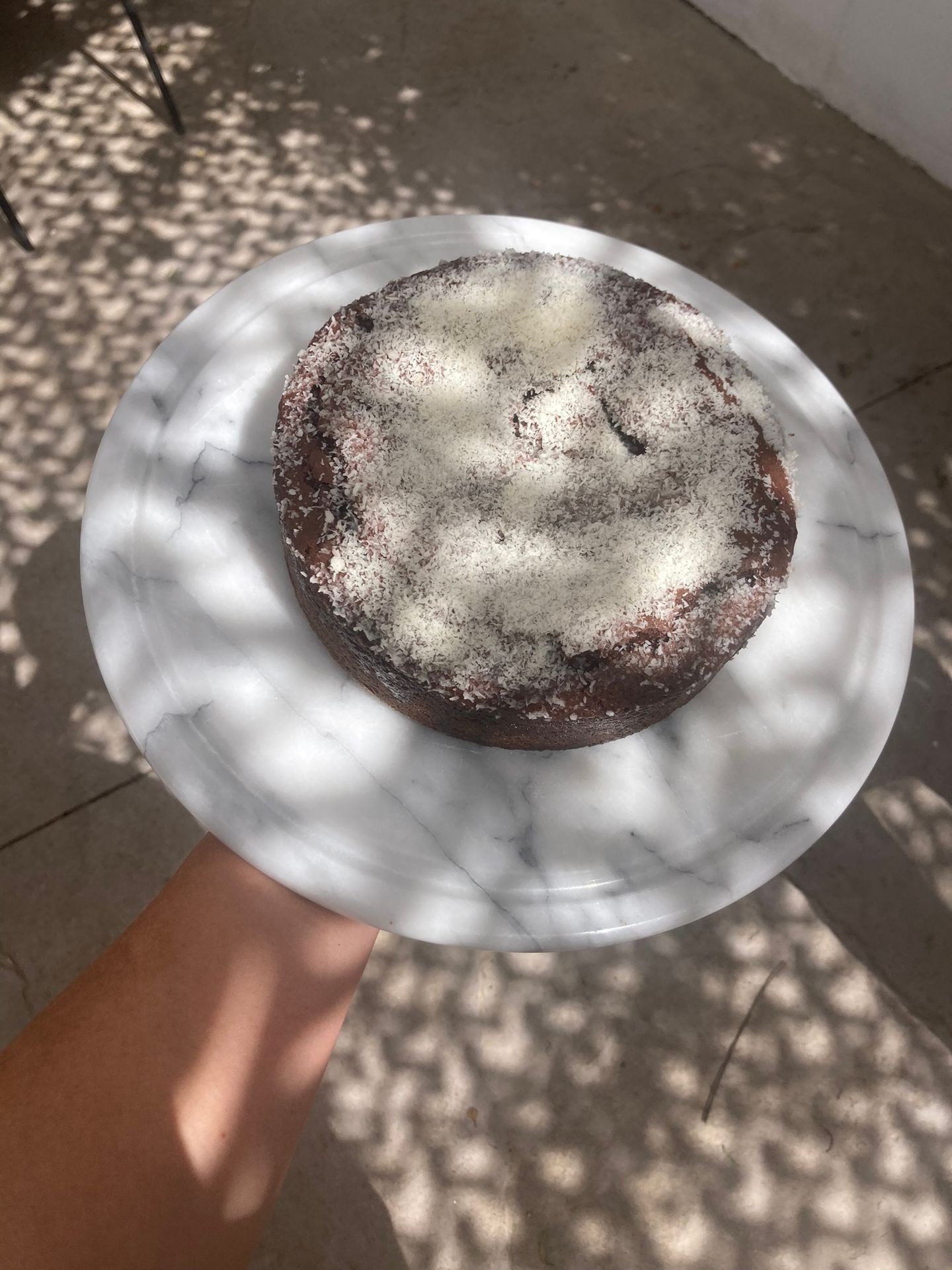 Hand holding a chocolate cake with powdered sugar on a marble plate, under dappled sunlight.
