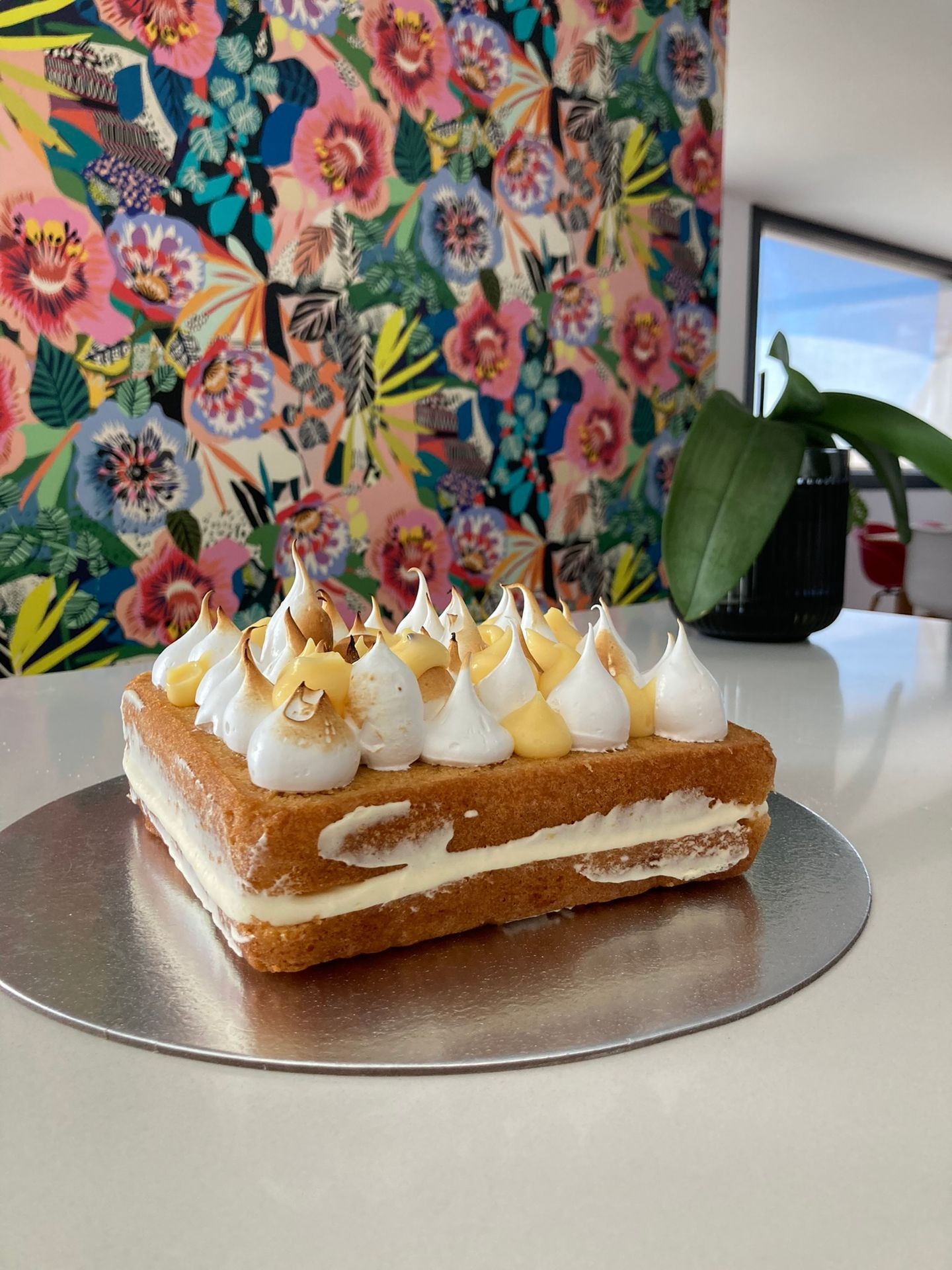 Rectangular cake with cream and meringue on a silver board, floral wallpaper background.