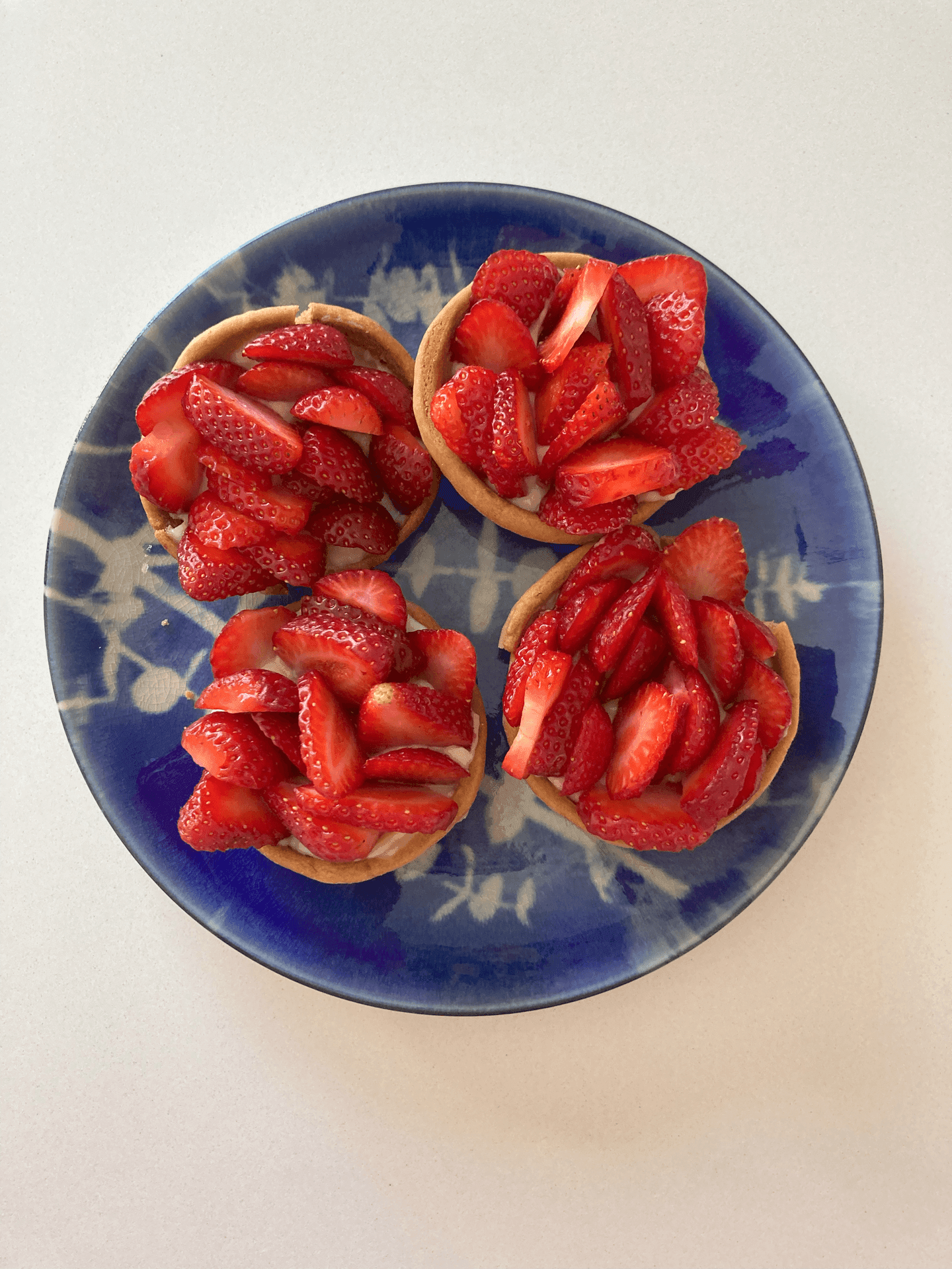 Four strawberry tarts on a blue and white ceramic plate, each topped with fresh strawberry slices.