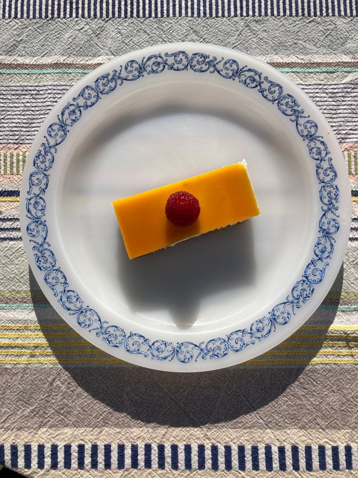 Slice of dessert topped with a raspberry on a decorated plate, placed on a patterned tablecloth.
