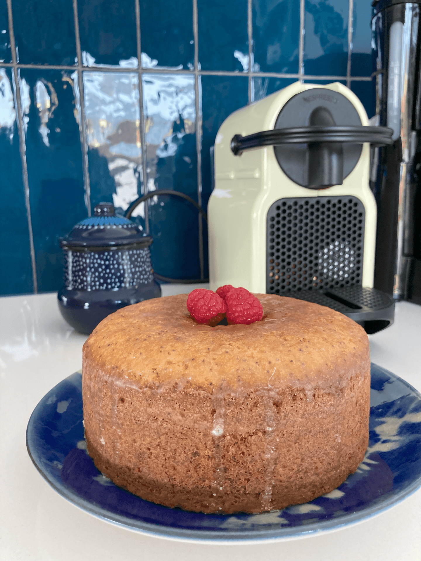 Round cake with raspberries on top, beside a coffee machine, set against blue tiled wall.